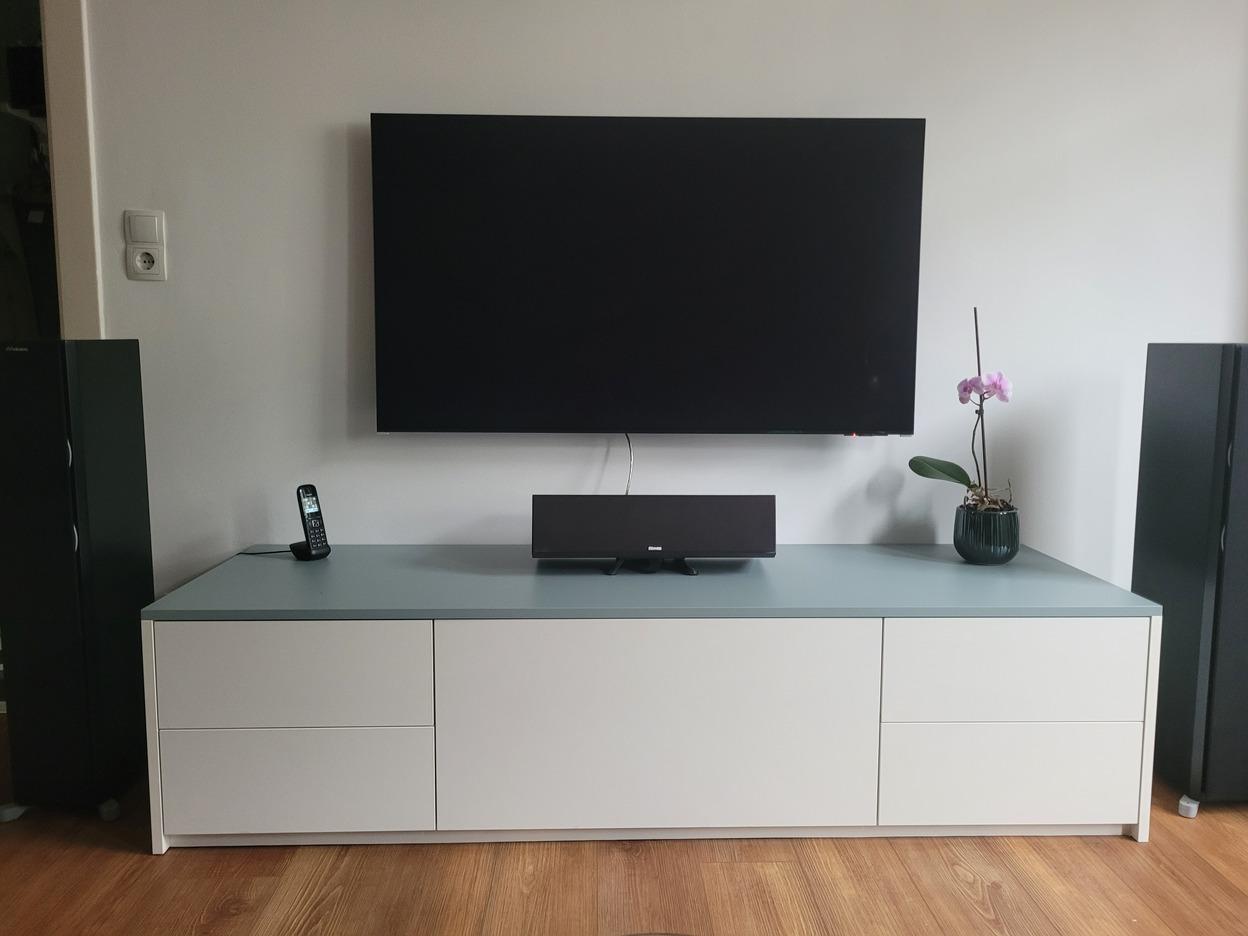 Modernes Wohnzimmer mit TV und Lowboard Ein modernes Wohnzimmer mit einem an der Wand montierten Fernseher und einem weißen Lowboard. Auf dem Lowboard befinden sich ein Telefon, eine Lautsprecherbox und eine blühende Orchidee.