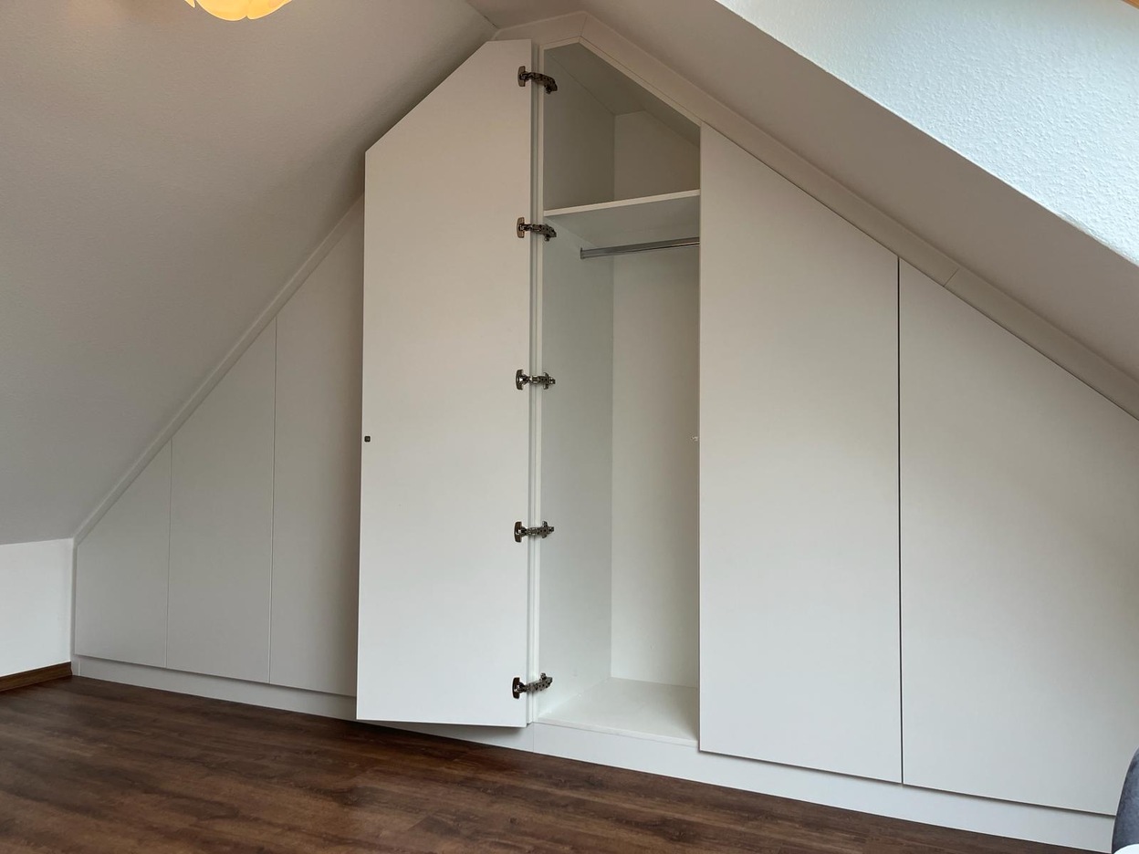 Ein nach Maß angefertigter Dachschrägenschrank in Weiß, integriert sich nahtlos in einen Raum mit Holzboden.