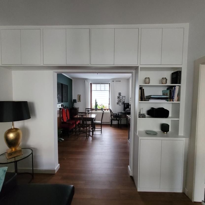 Ein modernes Wohnzimmer mit integriertem Essbereich. Der Raum ist mit weißen Einbauschränken und einem dunklen Lampenständer mit goldenem Lampenschirm ausgestattet. Der Boden ist aus Holz, und im Hintergrund befindet sich ein Fenster mit Vorhängen.