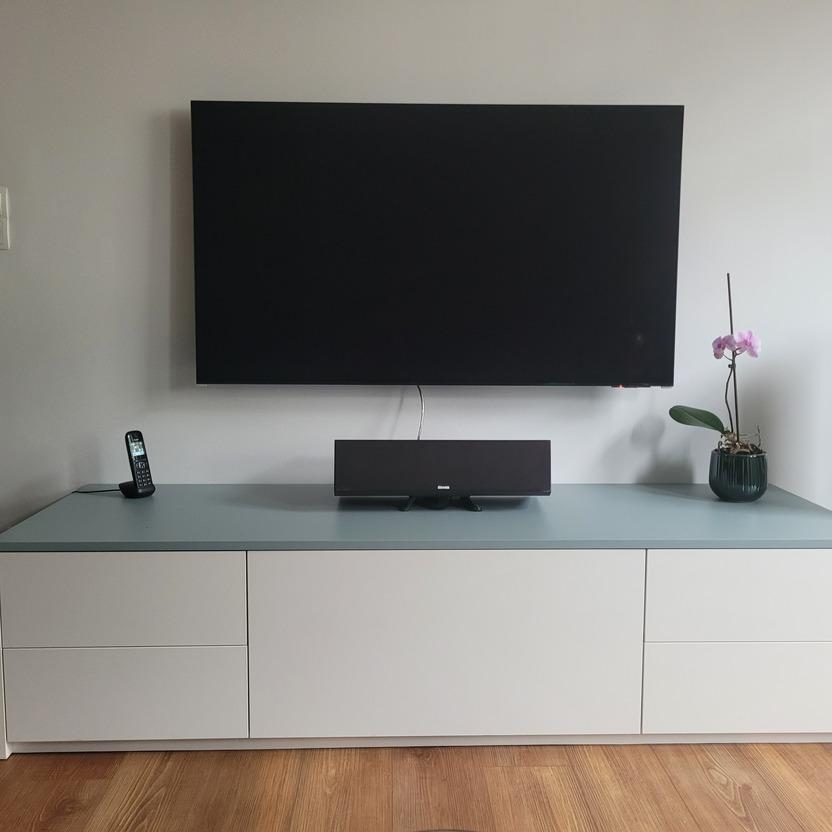 Ein modernes Wohnzimmer mit einem an der Wand montierten Fernseher und einem weißen Lowboard. Auf dem Lowboard befinden sich ein Telefon, eine Lautsprecherbox und eine blühende Orchidee.