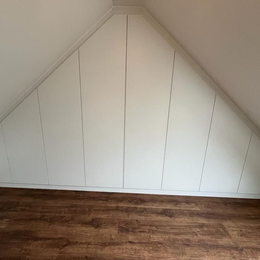 Ein Dachzimmer mit schräger Decke, ausgestattet mit weißen Einbauschränken, die sich perfekt an die Form des Raumes anpassen. Der Boden ist aus dunklem Holz.