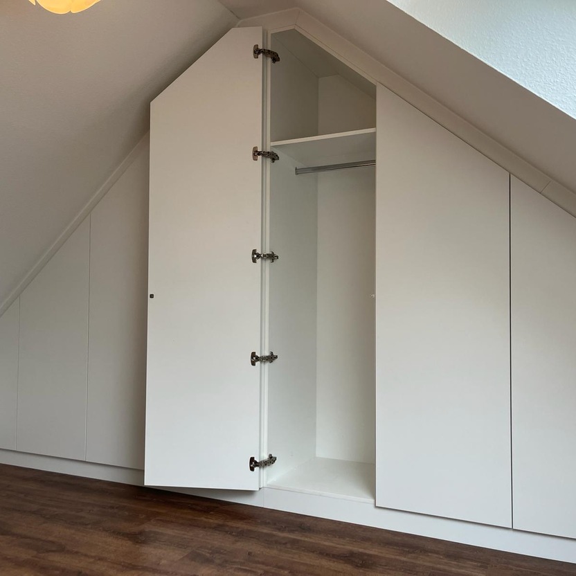 Ein nach Maß angefertigter Dachschrägenschrank in Weiß, integriert sich nahtlos in einen Raum mit Holzboden.