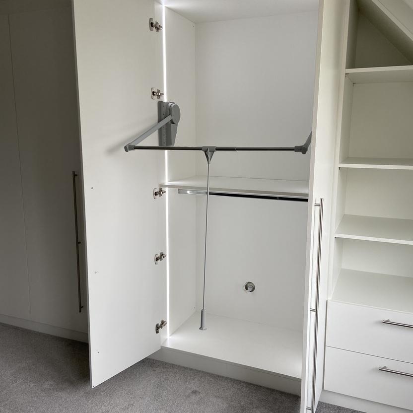 Ein geöffneter weißer Kleiderschrank in einem Dachzimmer mit schräger Decke. Der Schrank zeigt eine Kleiderstange und Regale. Der Boden ist mit grauem Teppichboden bedeckt.