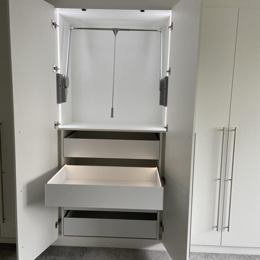 Innenausbau eines weißen Kleiderschranks, geöffnet, mit einem ausziehbaren Kleiderständer, zwei Einlegeböden und zwei Schubladen auf einem grauen Teppichboden.