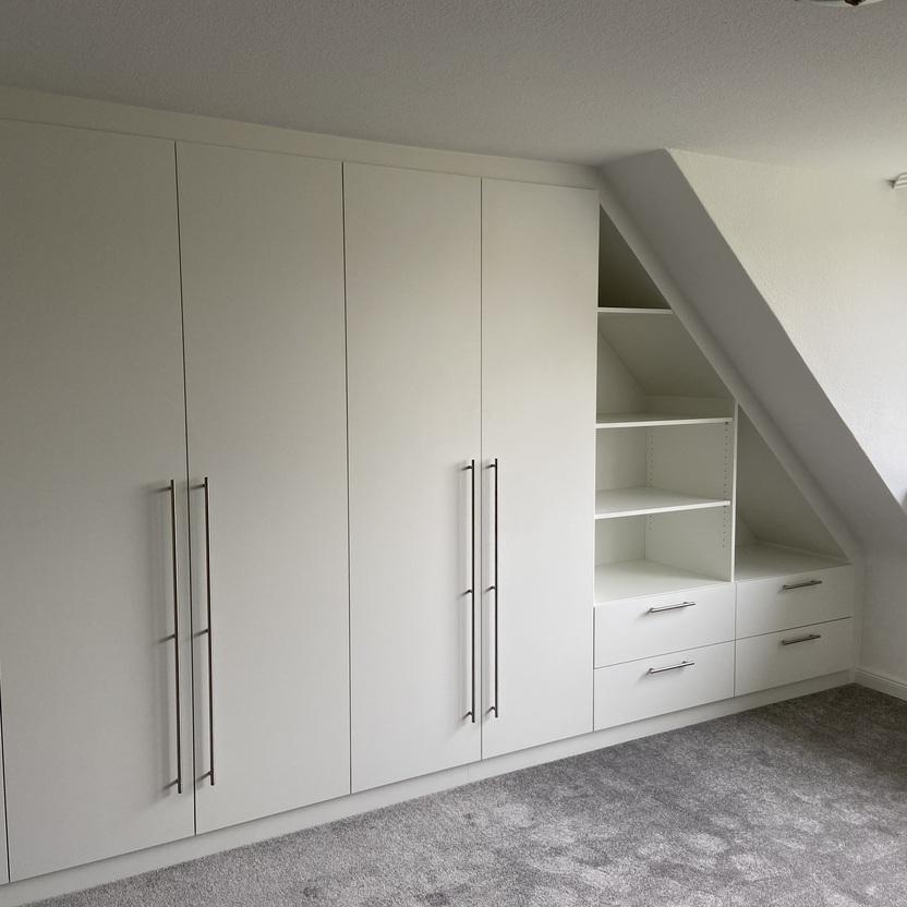 Weißer, maßgefertigter Innenausbau eines Schrankes unter einer Dachschräge mit mehreren Türen und Griffen, Schubladen und offenen Regalen neben einem Fenster.