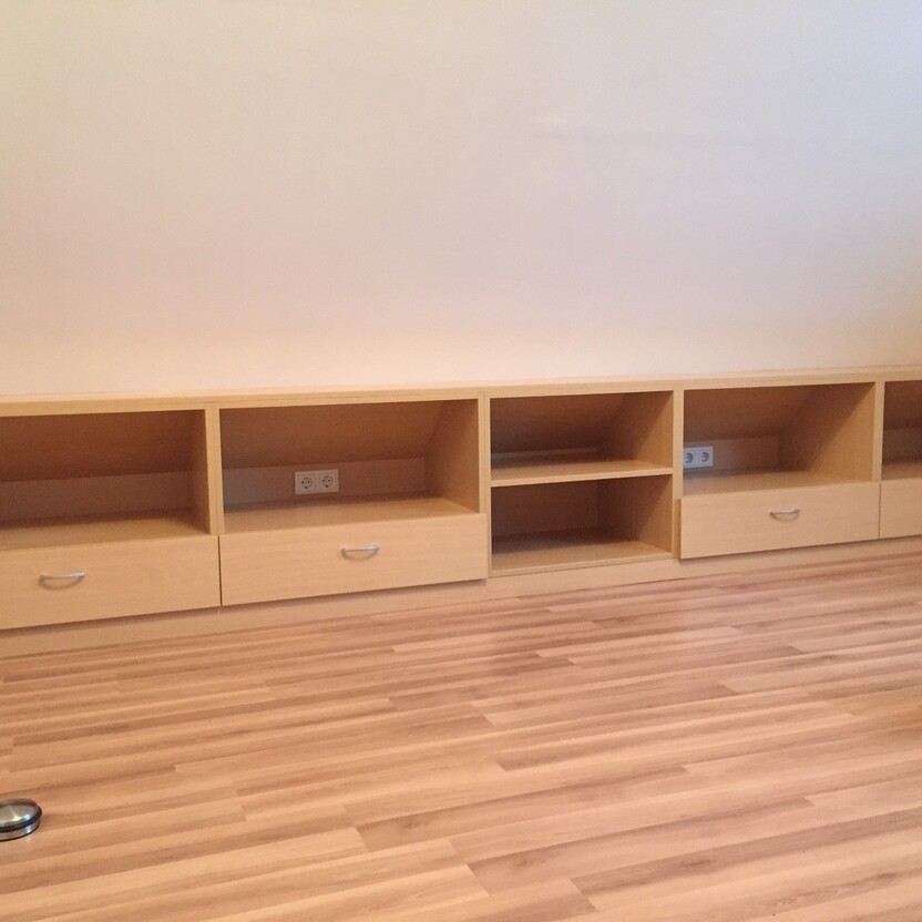 Maßgefertigte Holzmöbel als Teil des Innenausbaus unter einer Dachschräge, bestehend aus offenen Fächern und Schubladen auf Parkettboden.