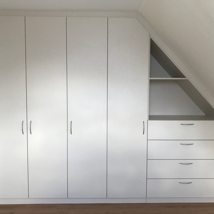 Moderner Innenausbau eines weißen Einbauschranks mit Türen und Schubladen, angepasst an eine Dachschräge, in einem hellen Raum mit Holzboden.