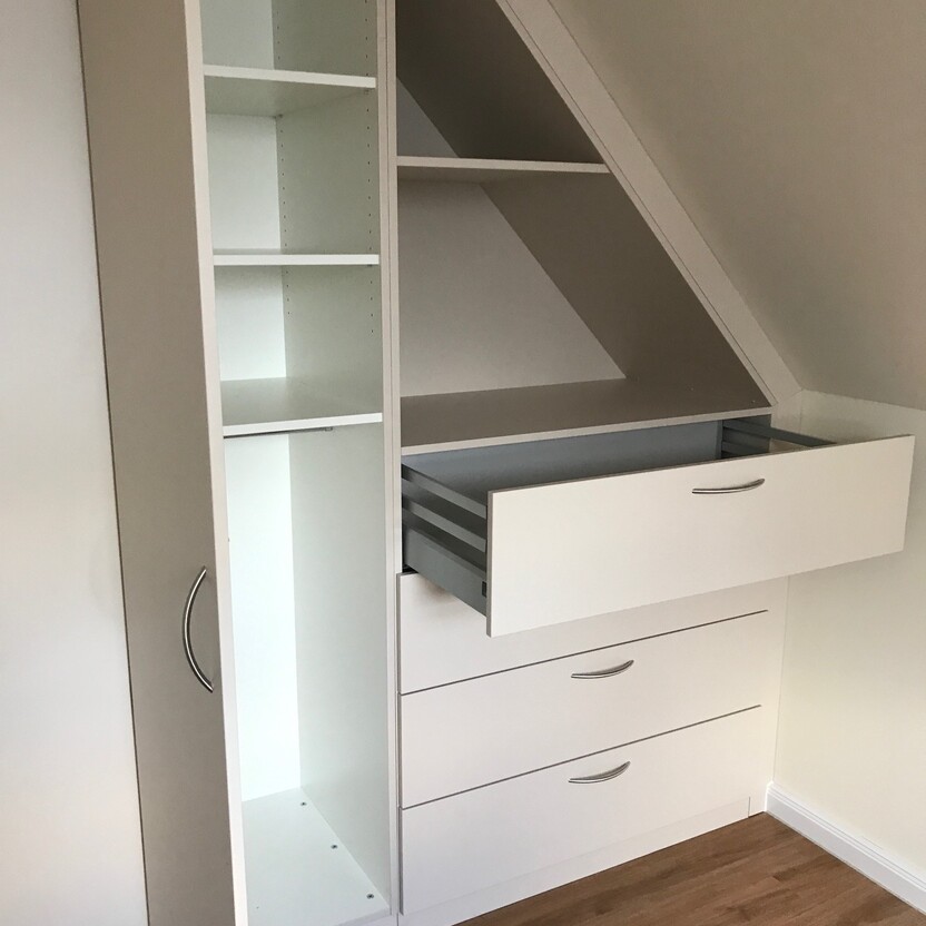 Teilansicht eines weißen Schranks als Innenausbau unter einer Dachschräge mit offenen Regalen, Schubladen und einem ausziehbaren Fach.
