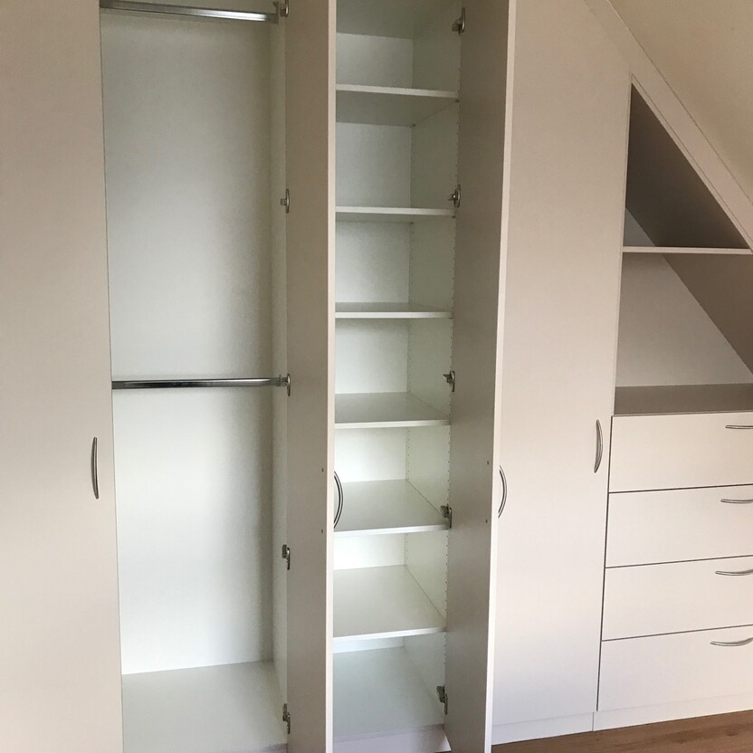 Innenausbau eines weißen Schrankes mit geöffneten Türen, offenbarend eine Kleiderstange und mehrere Regale, angepasst unter einer Dachschräge.