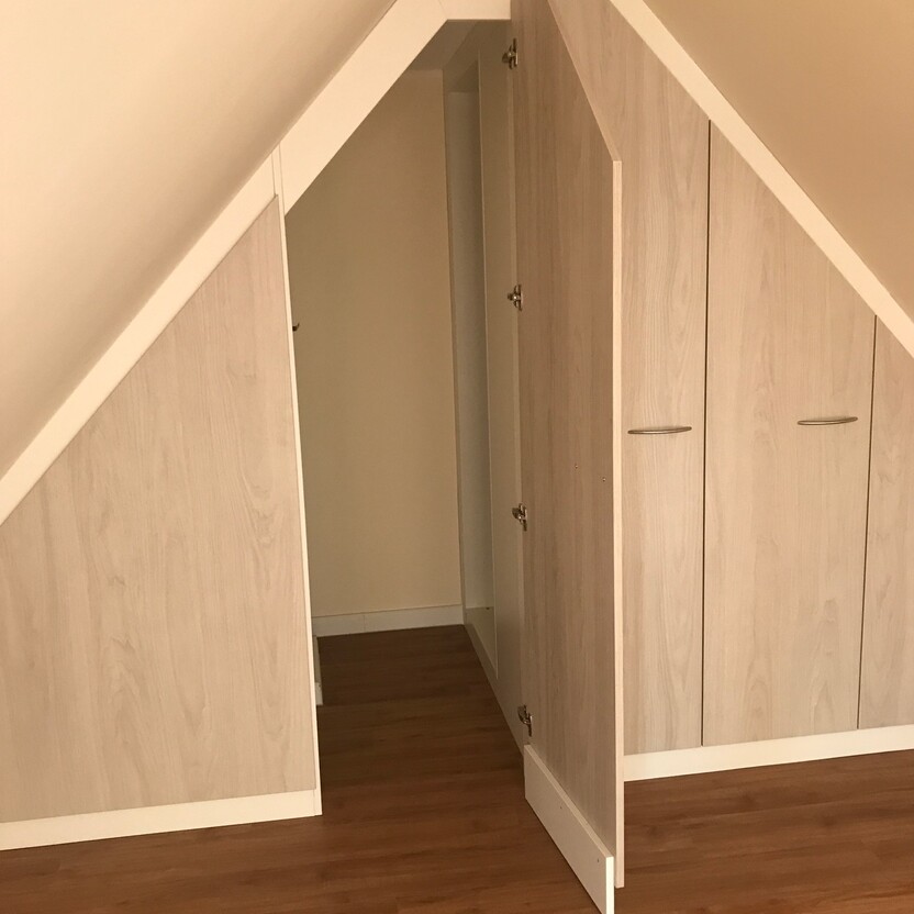 Holzfarbene Schränke als Teil des Innenausbaus in einem Dachzimmer, angepasst an die Dachschräge, mit sichtbarem Eingangsbereich.