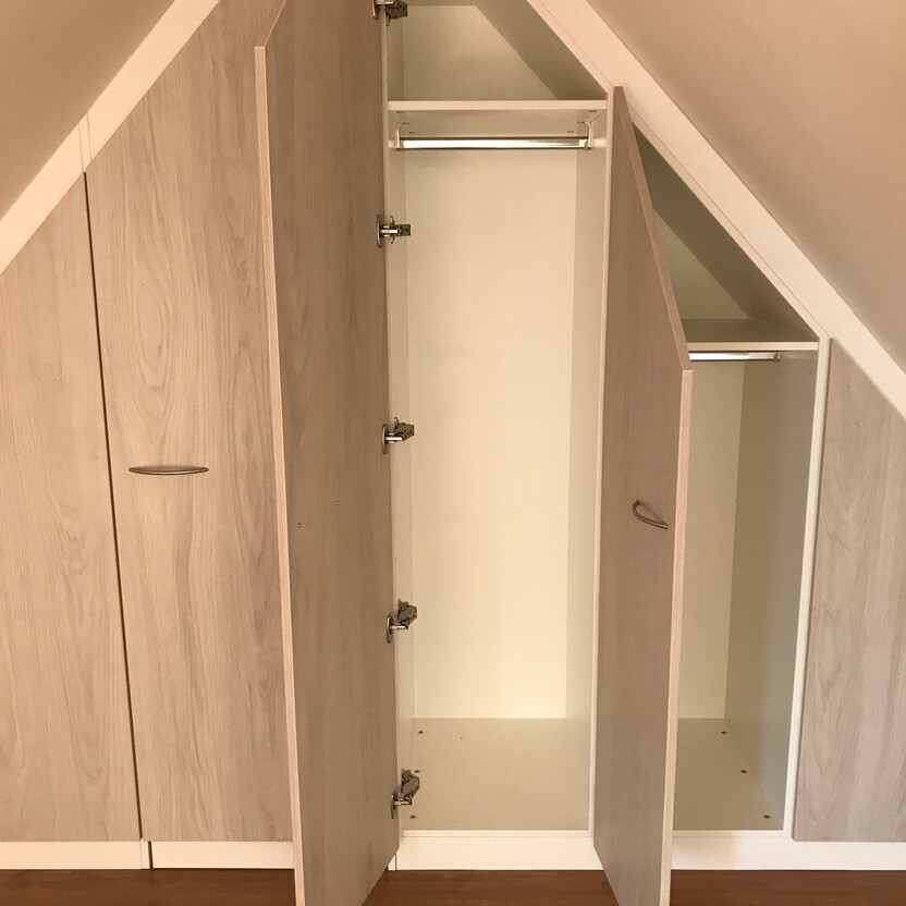 Innenausbau eines offenen, holzfarbenen Einbauschranks in einem Dachzimmer mit einer Kleiderstange und angrenzenden Regalen.