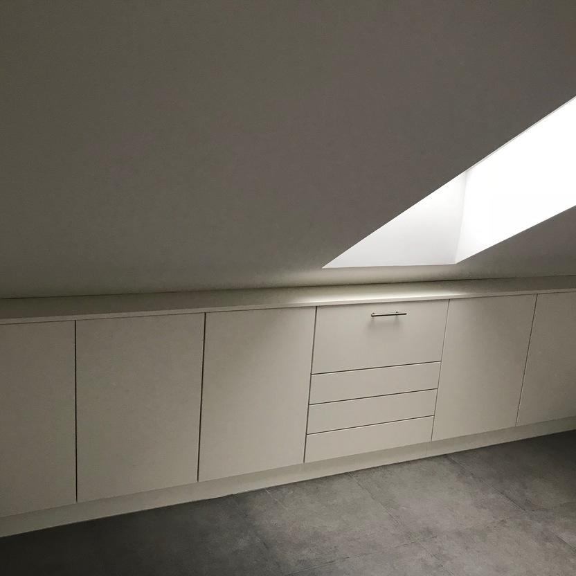 Weißer, schlichter Innenausbau eines Einbauschranks unter einer Dachschräge mit Dachfenster, der Stauraum und Schubladen bietet.