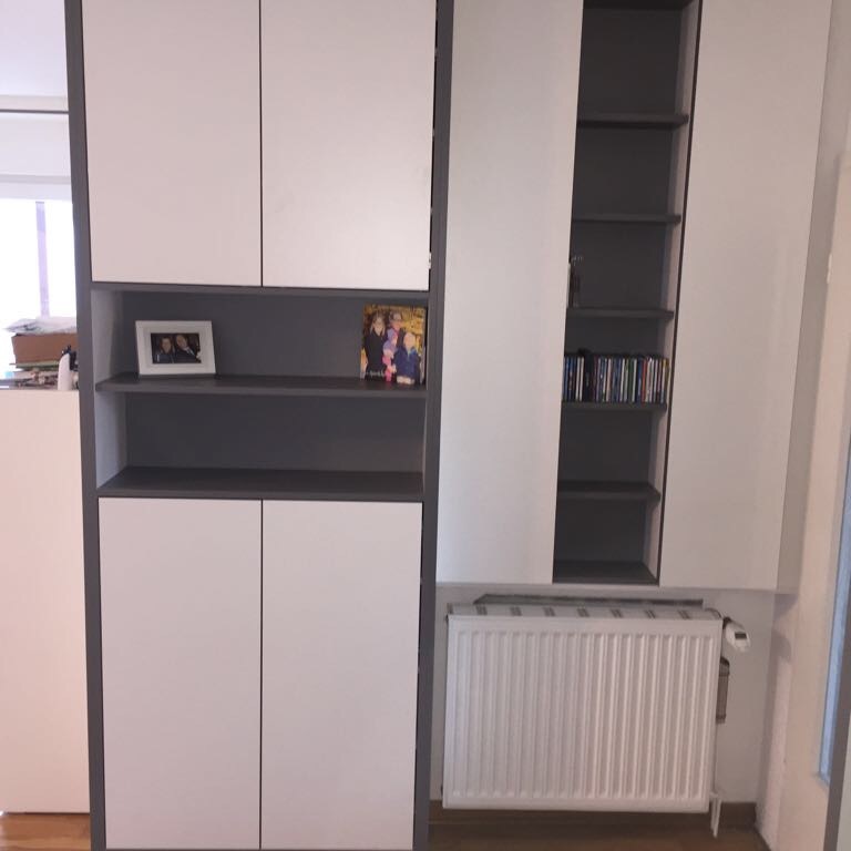 Weiß-grauer Einbauschrank mit Regalflächen und integriertem Platz für einen Heizkörper in einem modernen Innenausbau. Auf den Regalen stehen Bilderrahmen und CDs.