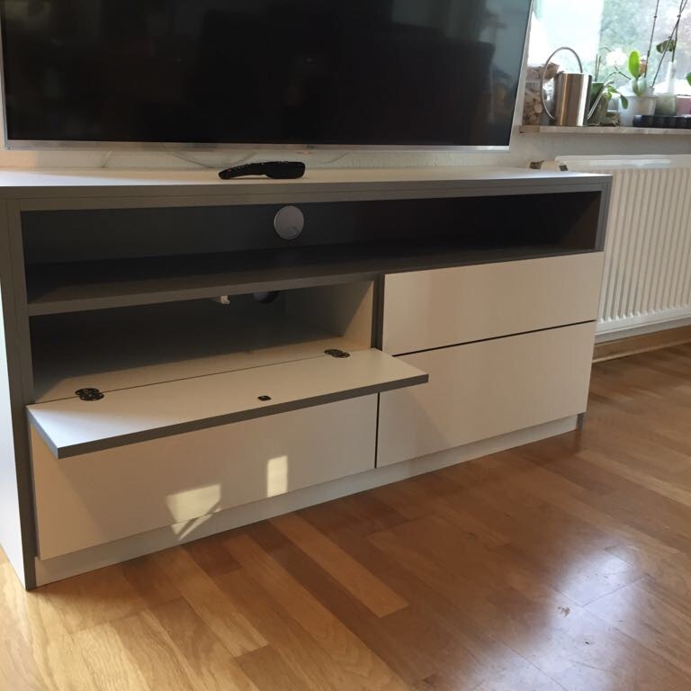 Weiß-graues TV-Möbel mit offenen Fächern und Schubladen in einem modernen Innenausbau. Auf dem Möbel steht ein Fernseher, daneben Pflanzen auf der Fensterbank.