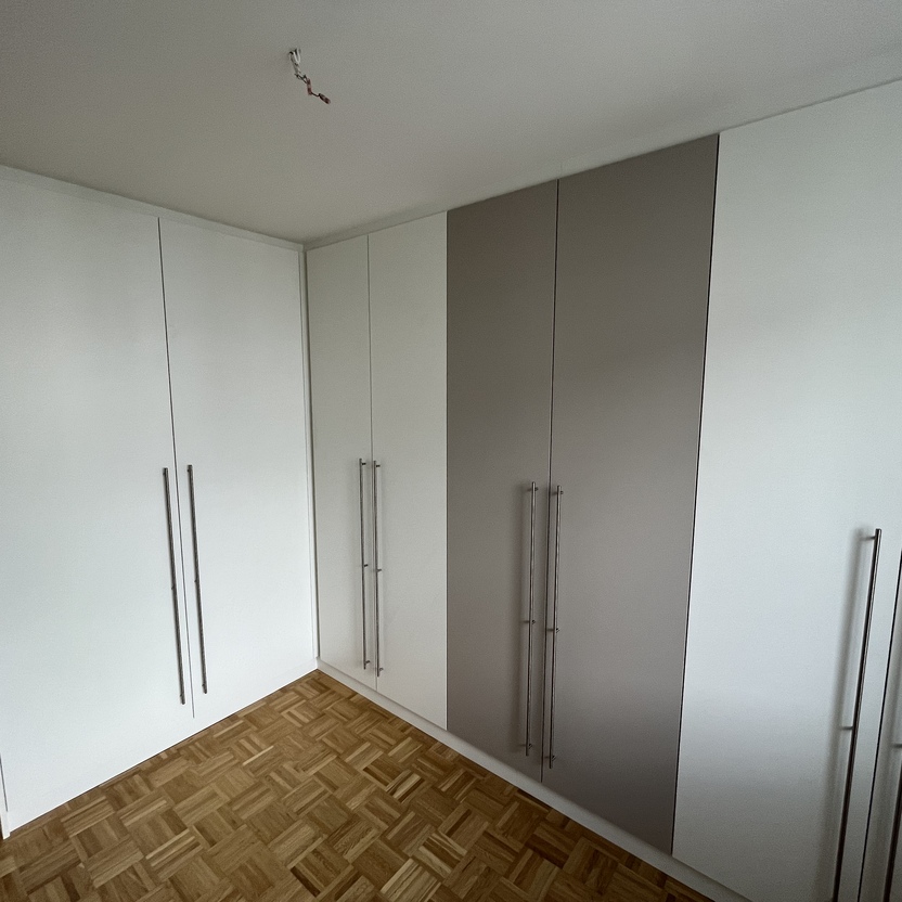 Großer, weiß-grauer Einbauschrank mit langen Griffen und Parkettboden in einem modernen Innenausbau. Der Schrank füllt die Ecke des Raumes aus und hat eine schlichte, elegante Optik.