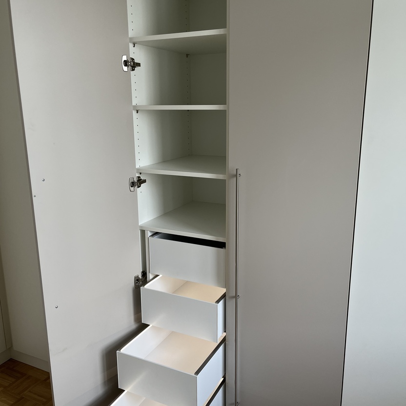 Geöffneter weißer Einbauschrank mit mehreren Regalen und vier ausziehbaren Schubladen in einem modernen Innenausbau. Der Schrank hat lange Griffe und steht auf einem Parkettboden.