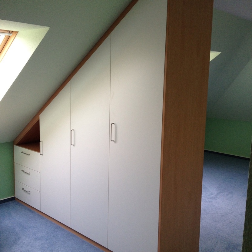 Maßgefertigter Einbauschrank in Holzoptik mit weißen Türen und Schubladen unter einer Dachschräge in einem modernen Innenausbau. Der Schrank passt sich perfekt der Schräge an und nutzt den Raum optimal aus.