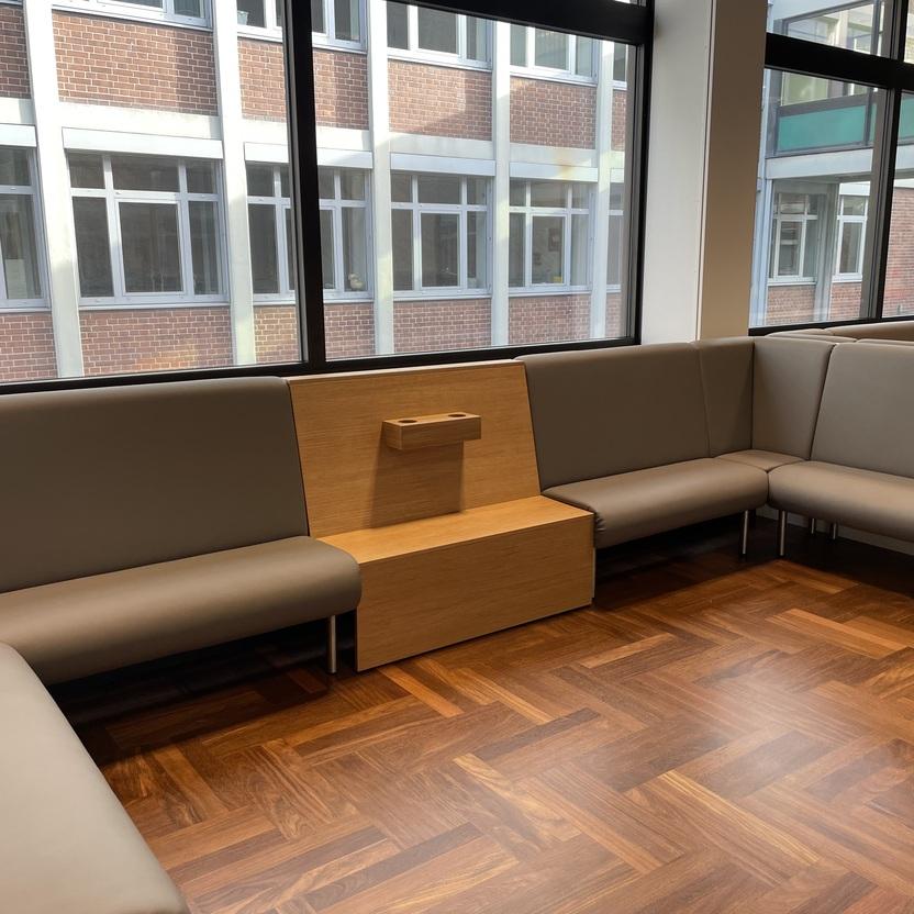 Moderner Warteraum mit einem grauen Ecksofa und einem hölzernen Zwischenelement in einem hochwertigen Innenausbau. Der Raum hat große Fenster und einen Boden in Holzoptik.