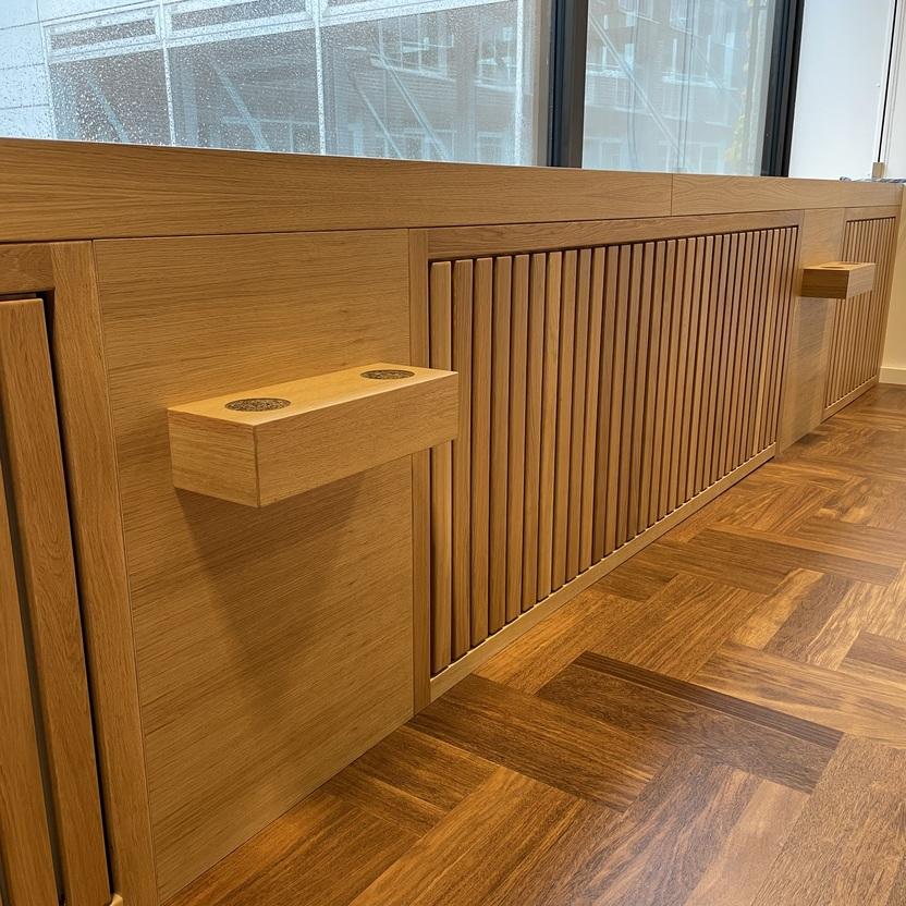 Detaillierte Ansicht von Holzelementen mit vertikalen Lamellen und integrierten Ablagen in einem modernen Innenausbau. Der Raum hat große Fenster und einen eleganten Holzboden.