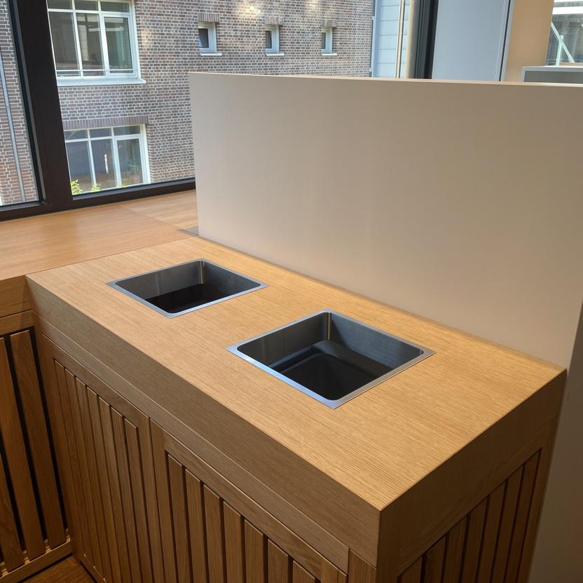 Modernes Abfalltrennsystem aus Holz mit zwei Einwurfschächten in einem hochwertigen Innenausbau. Das System steht vor großen Fenstern, die einen Blick auf ein Backsteingebäude bieten.