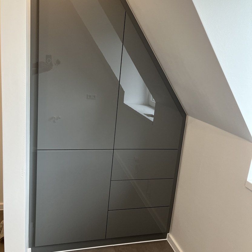 Maßgefertigter, grauer Einbauschrank unter einer Dachschräge in einem modernen Innenausbau. Der Schrank hat eine glatte Oberfläche und bietet sowohl Schubladen als auch Stauraum hinter Türen.