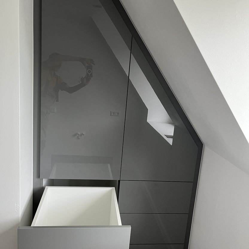 Geöffneter, grauer Einbauschrank unter einer Dachschräge in einem modernen Innenausbau. Der Schrank verfügt über ausziehbare Schubladen und bietet optimalen Stauraum in einem kleinen Raum.