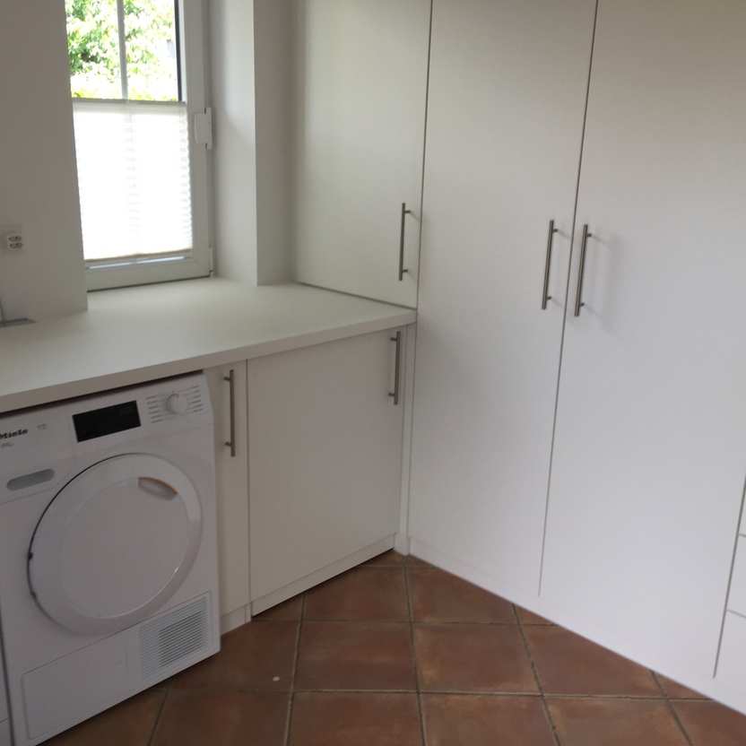 Moderner Hauswirtschaftsraum mit weißen Einbauschränken, einer Arbeitsplatte, Waschmaschine und Trockner in einem hochwertigen Innenausbau. Der Raum hat ein Fenster und Fliesenboden.