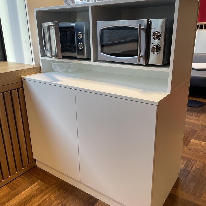 Weißes Küchenmöbel mit zwei integrierten Mikrowellen in einem modernen Innenausbau. Das Möbelstück steht auf einem eleganten Holzboden und vor großen Fenstern, die viel Licht hereinlassen.