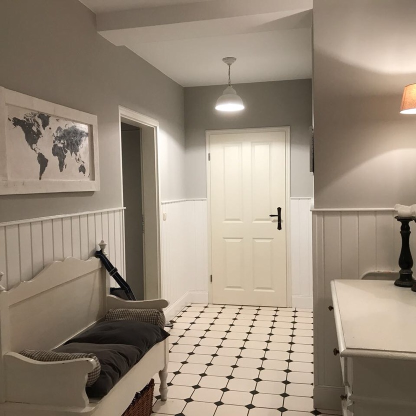 Eleganter Flur mit weißer Sitzbank, Wanddekoration, und schwarzen Leuchten in einem stilvollen Innenausbau. Der Flur hat eine helle, halbhoch getäfelte Wand und einen schwarz-weißen Fliesenboden.