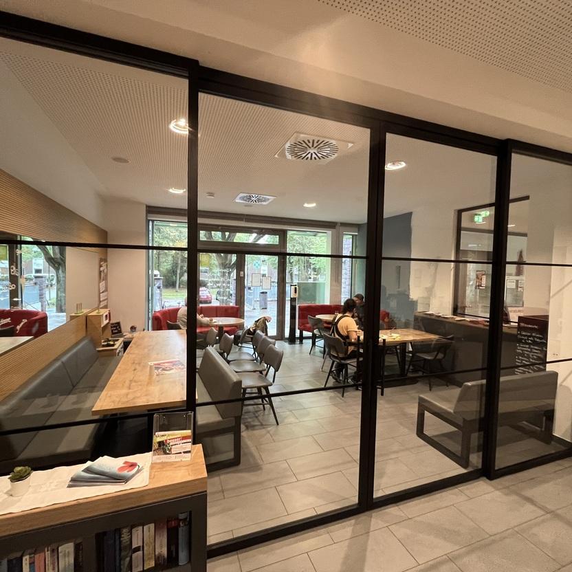 Moderner Besprechungsraum mit Glaswänden, großem Holztisch und grauen Sitzbänken in einem stilvollen Innenausbau. Der Raum hat große Fenster, die viel Tageslicht hereinlassen.