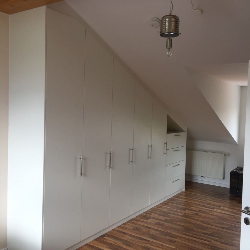 Weißer maßgefertigter Einbauschrank mit langen Griffen und integrierten Schubladen unter einer Dachschräge in einem modernen Innenausbau. Der Raum hat einen Holzfußboden und eine moderne Beleuchtung.