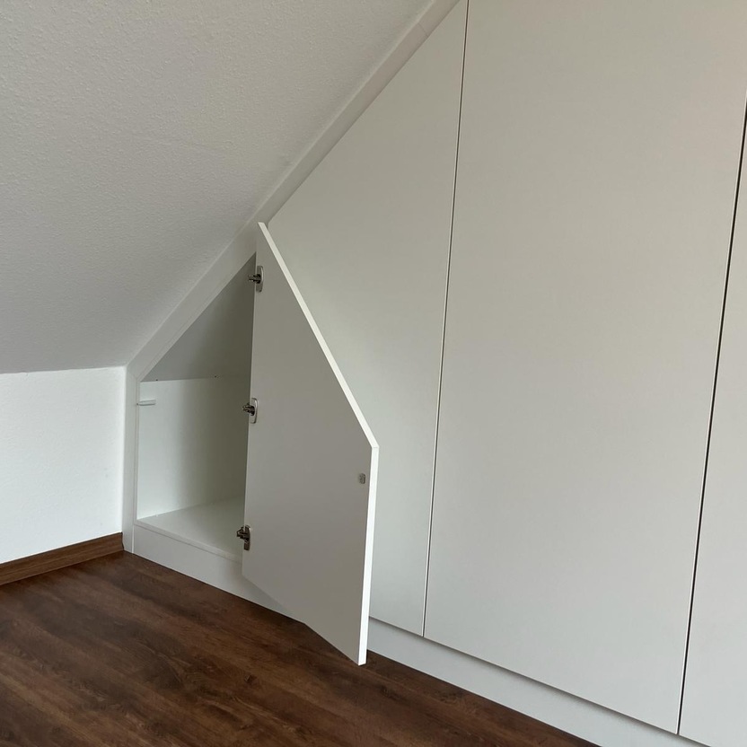 Ein Dachzimmer mit schräger Decke und einem weißen Einbauschrank. Eine Schranktür ist geöffnet und zeigt den Stauraum im Inneren. Der Boden ist aus dunklem Holz.