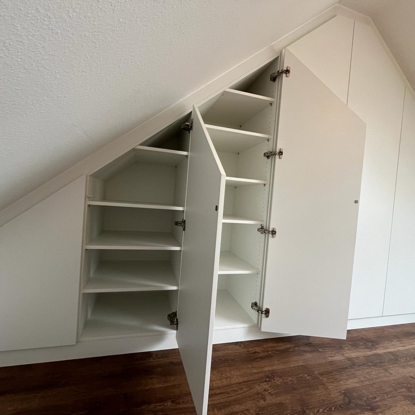 Ein Dachzimmer mit schräger Decke und weißen Einbauschränken. Mehrere Schranktüren sind geöffnet und zeigen die eingebauten Regale und Stauraum. Der Boden ist aus dunklem Holz.