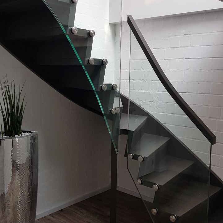 Schwarze Holztreppe mit Glasgeländer und Metallbeschlägen in einem hellen Treppenhaus mit weißen Wänden und großen Fenstern.