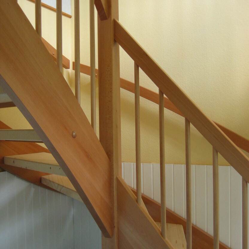 Klassische Holztreppe mit hellem Geländer und geschwungenem Handlauf aus Massivholz vor einer gelben Wand.