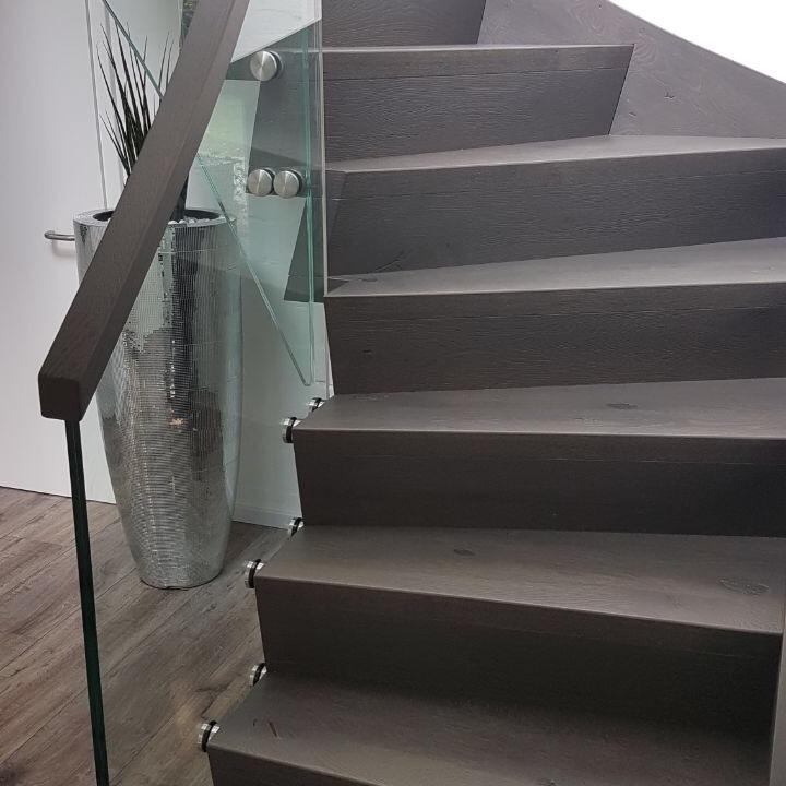 Moderne Holztreppe mit dunklen Stufen, Glasgeländer und Edelstahlbefestigungen in einem hellen Treppenhaus.