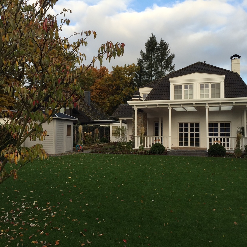 Helles Landhaus mit überdachter Holzveranda, grauem Ziegeldach und weitläufigem Garten mit Bäumen und Rasenfläche.