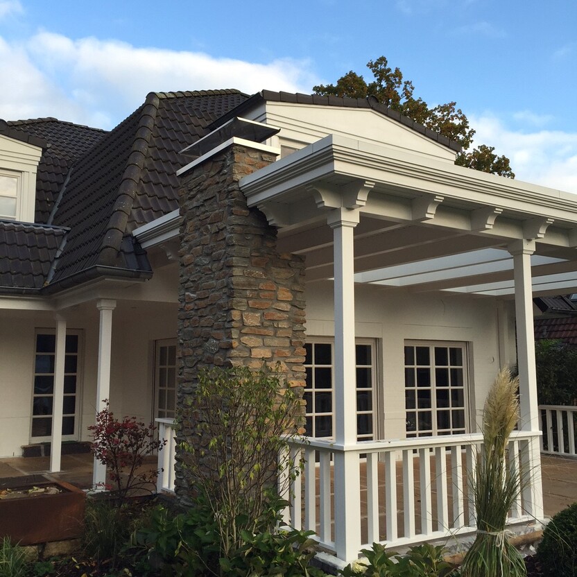 Überdachte Veranda mit weißem Holzgeländer und gemauertem Kamin, umgeben von Gartenpflanzen unter blauem Himmel.