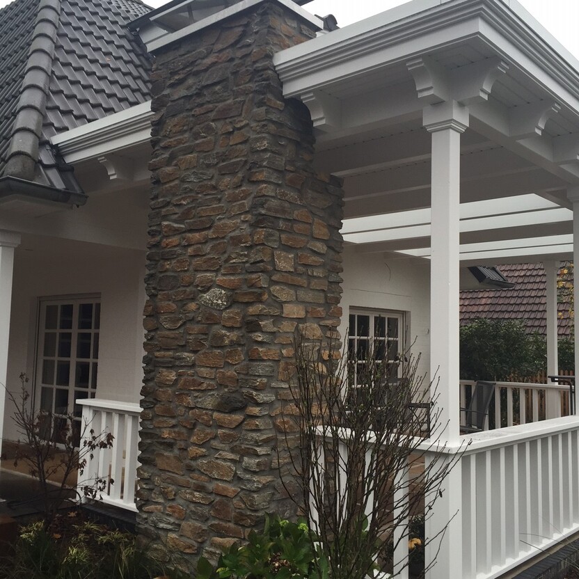 Steinsäule mit integriertem Kamin neben einer überdachten Holzveranda mit weißen Pfosten und Geländer.