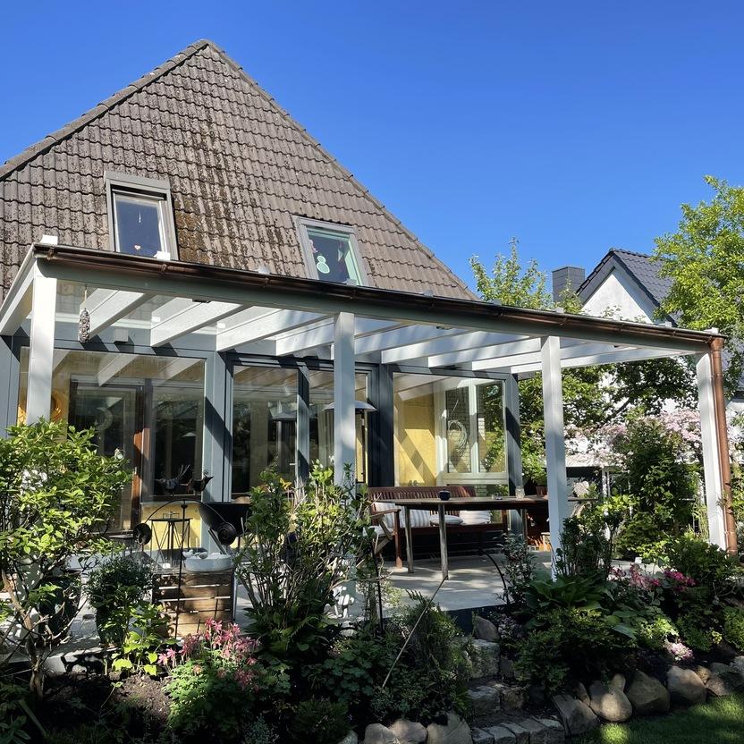 Helle Terrassenüberdachung aus weiß gestrichenem Holz mit Sitzbereich im Garten eines Einfamilienhauses.