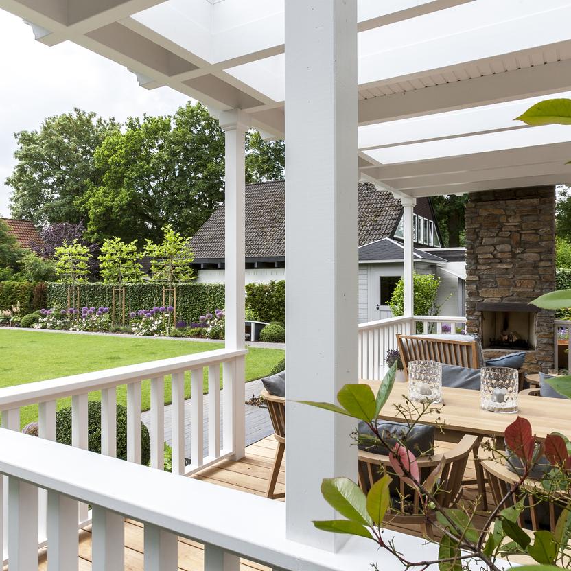 Holzveranda mit weißem Geländer, Blick auf gepflegten Garten und gemütlichen Essbereich mit Holztisch und Kamin.